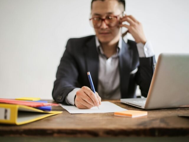 Businessman talking on phone while writing notes and using a laptop at desk.